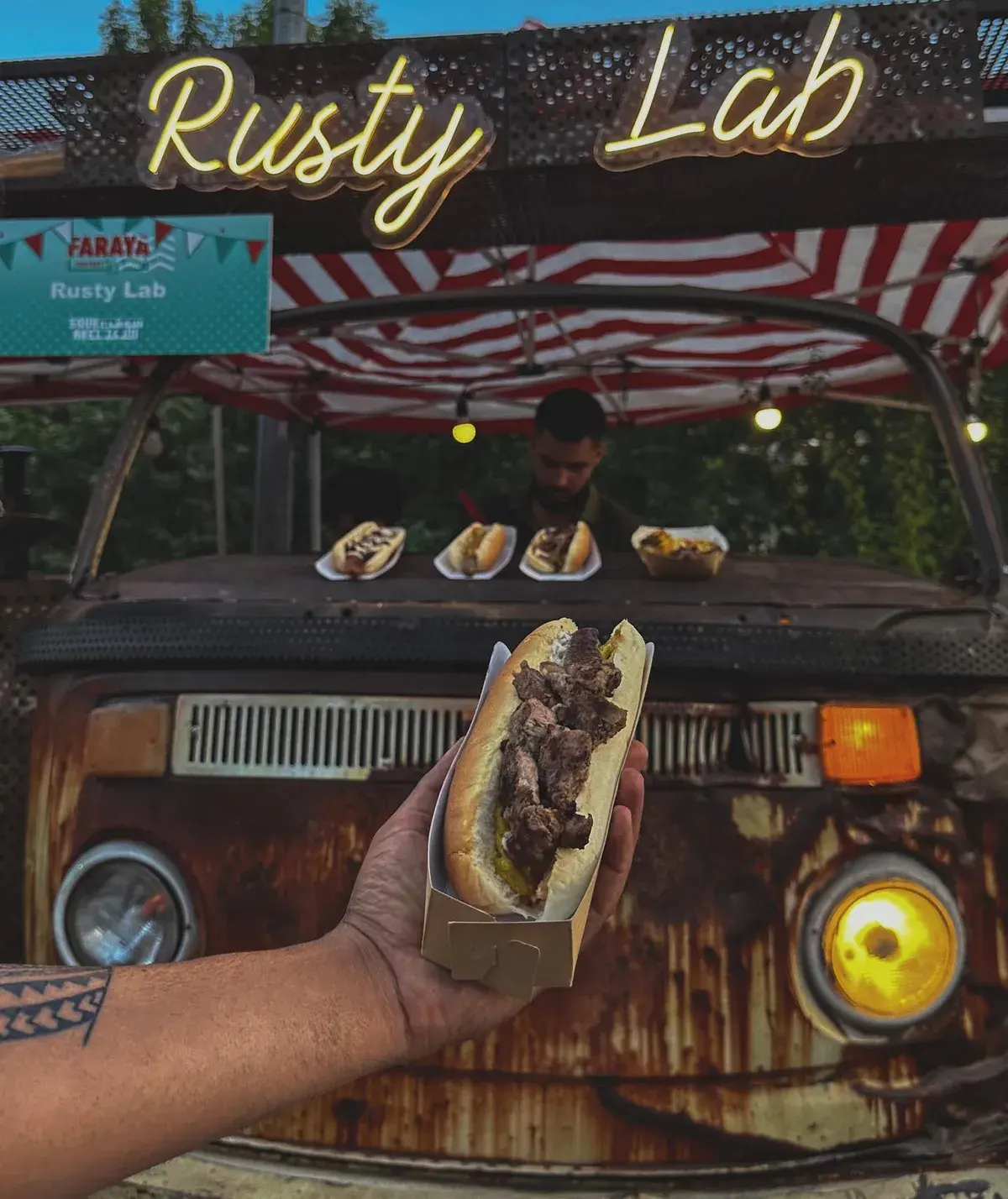 A steak sandwich held up in front of the rusty van's headlights, under the neon Rusty Lab sign