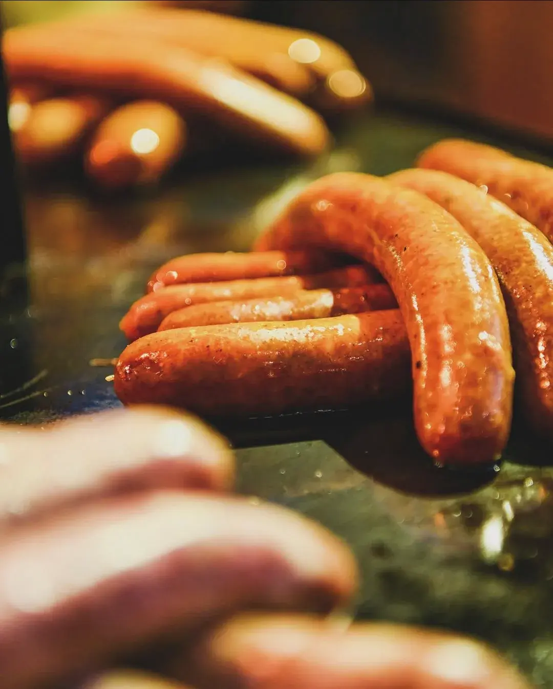 Close-up of glistening sausages browning on the flat-top grill, the next batch waiting behind