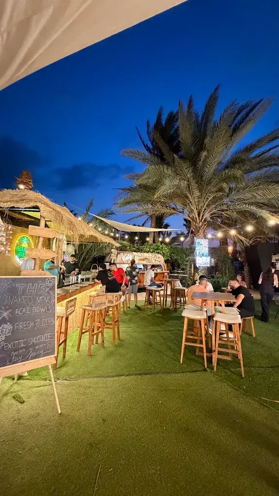 High tables and stools on the grass under palm trees and string lights at night, with the Rusty Lab van behind